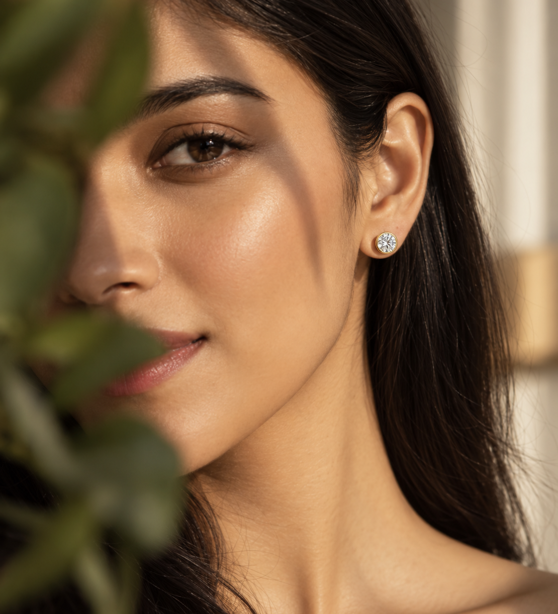 A close-up lifestyle shot of a woman with dark hair wearing a small, round gold stud earring with a clear gemstone, partially framed by soft-focus green leaves in the foreground.