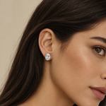 A close-up of a woman's ear wearing a small oval stud earring with a sparkling clear gemstone in a silver-toned setting, against a soft beige background.