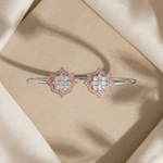 A pair of silver-toned bangle bracelets with diamond-like floral clusters accented by pink stones, displayed on a beige textured surface surrounded by draped cream satin fabric.