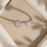 A pair of silver-toned bangle bracelets with diamond-like floral clusters accented by pink stones, displayed on a beige textured surface surrounded by draped cream satin fabric.