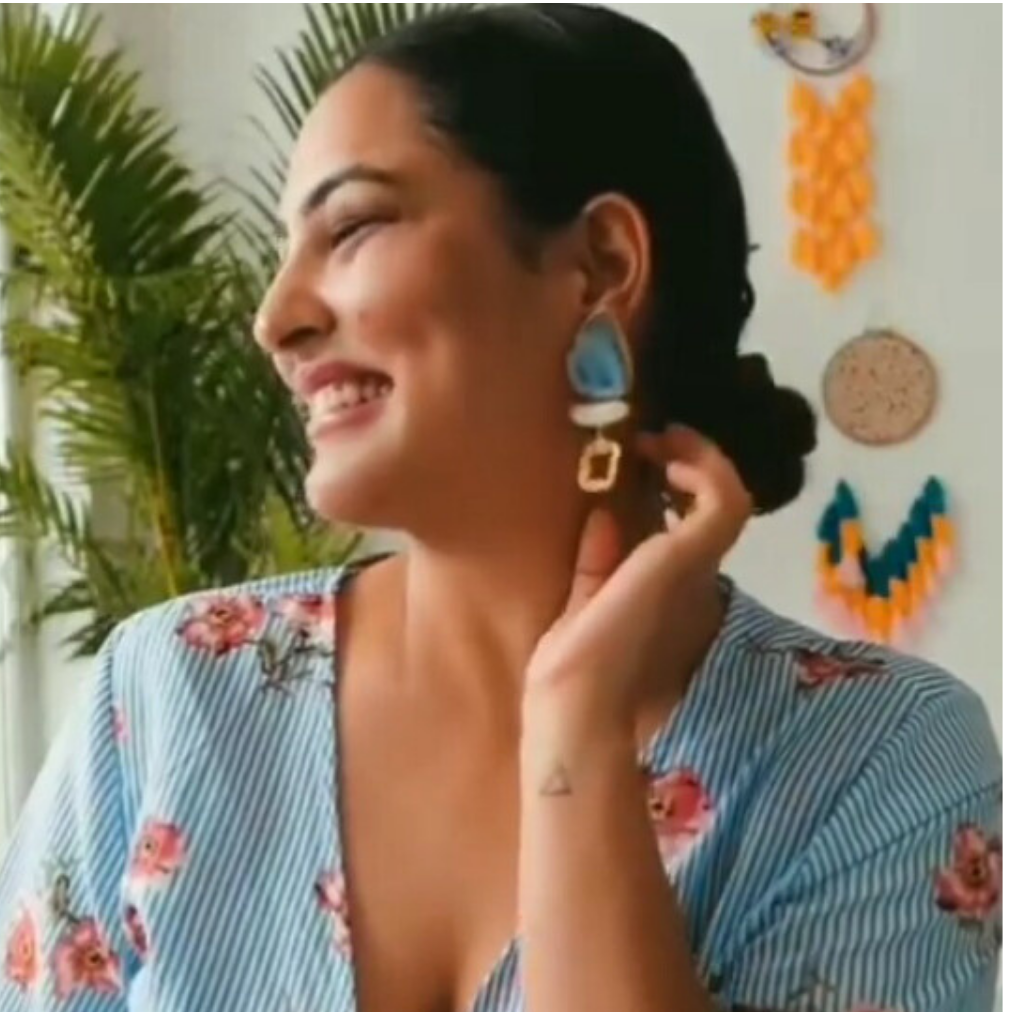 Woman wearing blue earrings with a floral top, indoors with plants and wall decorations.