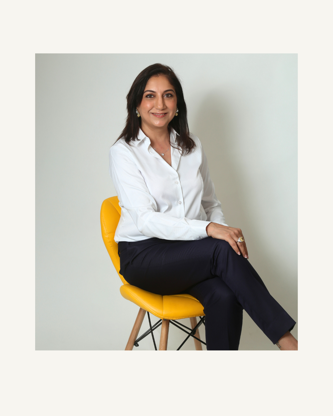 Woman sitting on a yellow chair wearing a white shirt and dark pants against a plain background