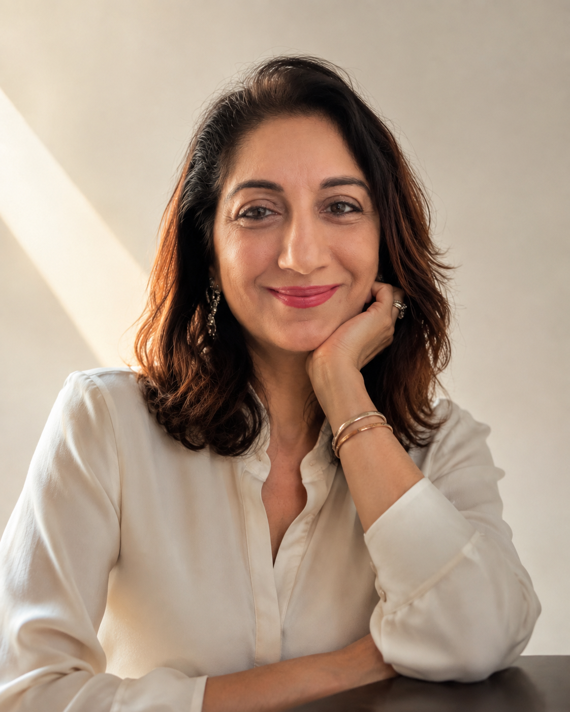 A woman with shoulder-length brown hair resting her chin on her hand, wearing a white button-up blouse and gold bracelets, set against a warm beige background with a diagonal light beam.