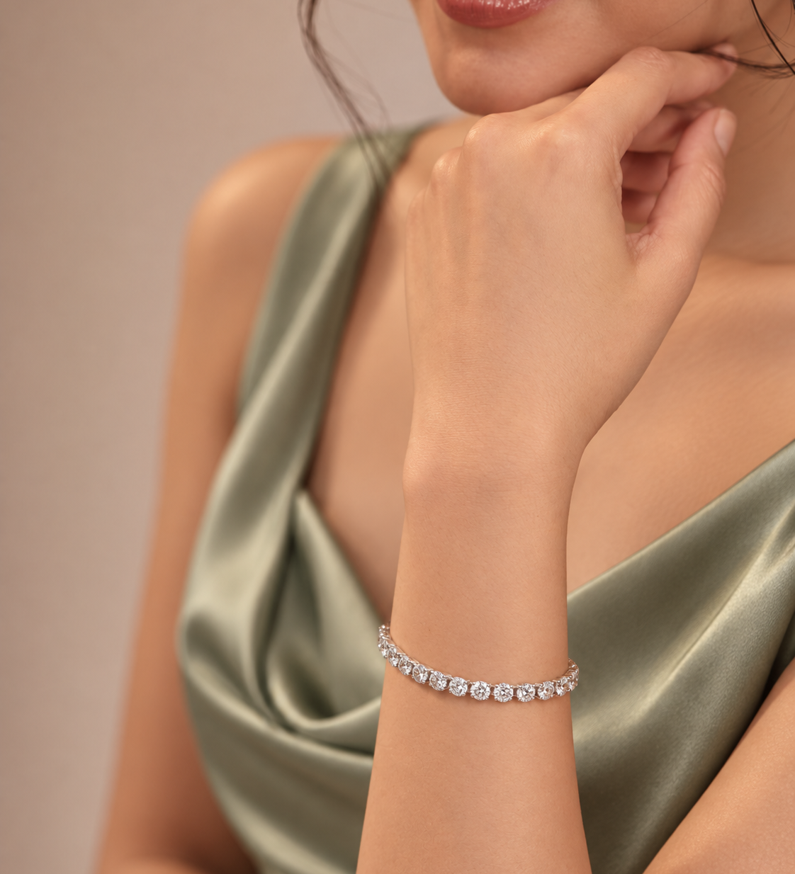 A close-up lifestyle shot of a model wearing a delicate silver tennis bracelet adorned with clear round-cut gemstones. The model is wearing a satin-look sage green dress against a soft, warm beige background.