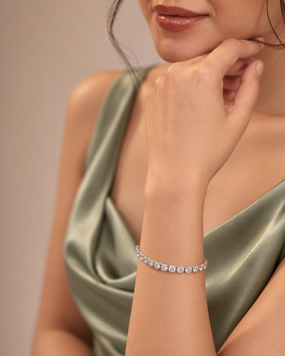 A close-up lifestyle shot of a model wearing a delicate silver tennis bracelet adorned with clear round-cut gemstones. The model is wearing a satin-look sage green dress against a soft, warm beige background.