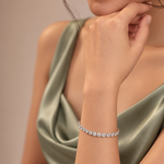 A close-up lifestyle shot of a model wearing a delicate silver tennis bracelet adorned with clear round-cut gemstones. The model is wearing a satin-look sage green dress against a soft, warm beige background.