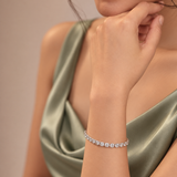 A close-up lifestyle shot of a model wearing a delicate silver tennis bracelet adorned with clear round-cut gemstones. The model is wearing a satin-look sage green dress against a soft, warm beige background.