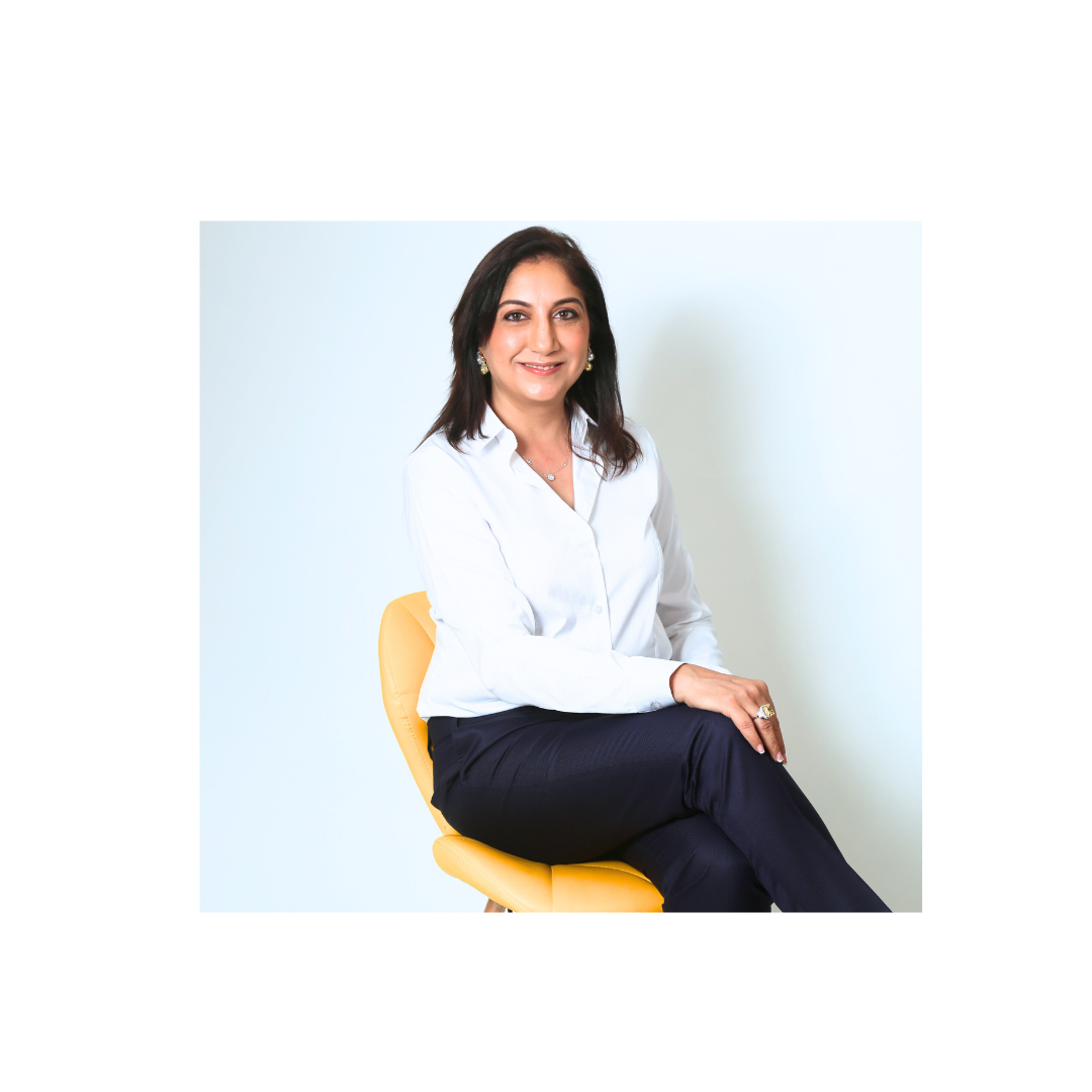 Shabnam Bhojwani - A confident woman in a white shirt and dark pants sits on a yellow modern chair against a light blue background, smiling at the camera.